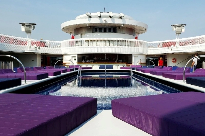 The image features a modern pool area on cruise ship. In the foreground, there are several purple lounge beds arranged around the water. The pool itself has a sleek design, reflecting the surroundings. In the background, an upper deck structure is visible with a rounded shape and large windows, contributing to a contemporary aesthetic. The sky is clear and blue, indicating good weather.